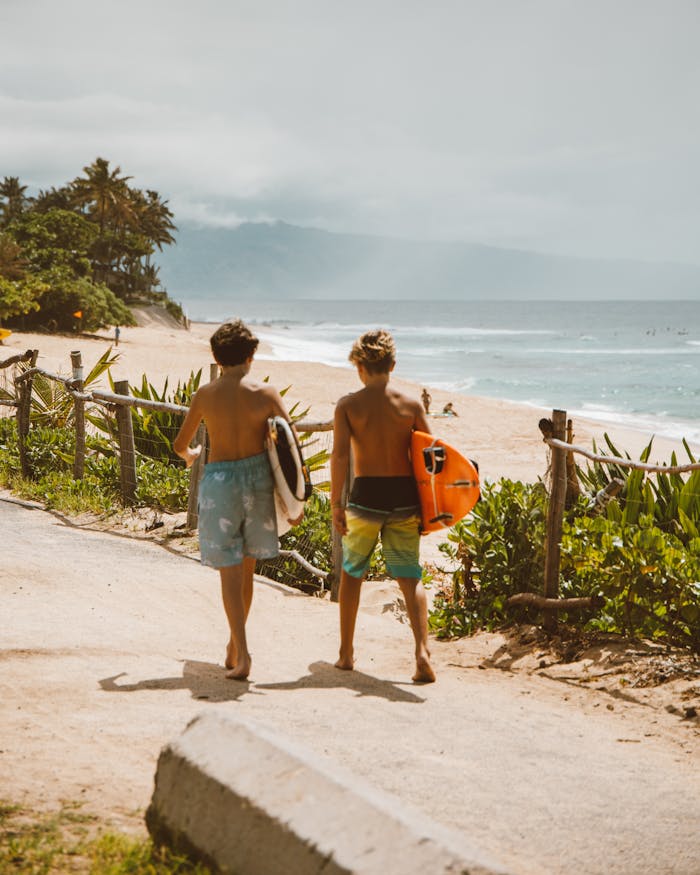 Destinations Two young surfers walk along the tropical beach path towards the ocean, carrying surfboards on a bright day.